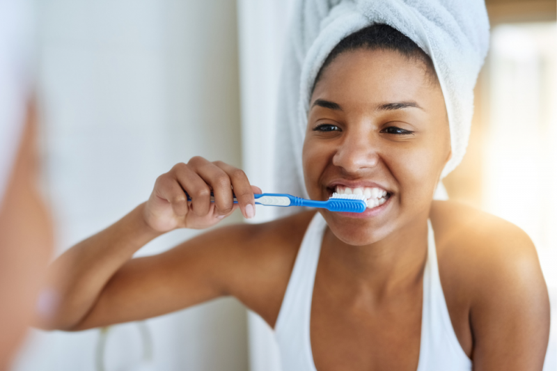 A young woman with a white towel wrapped around her hair looks in the mirror as she brushes her teeth