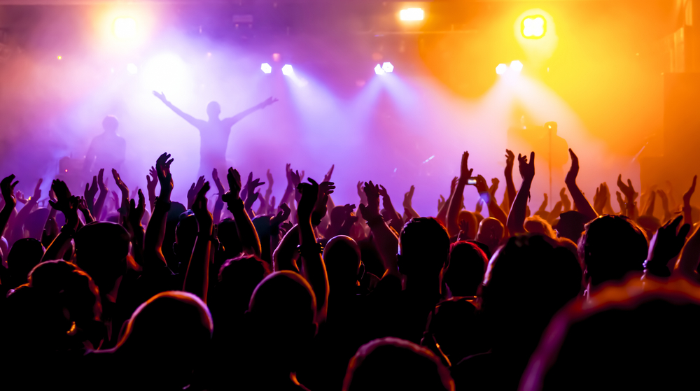 A large crowd stand in front of a musical festival stage, the silhouette of the band can be seen against smoke lit up pink and yellow from the stage lights. 