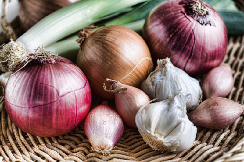A photograph of a basket containing garlic, brown and red onions, and spring onions