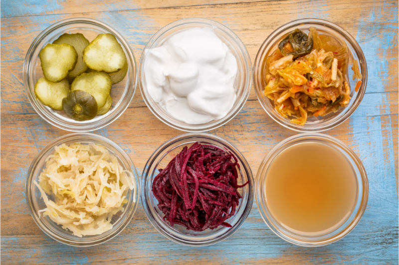 A range of fermented foods great for gut health - top view of small glass bowls against a rough wooden table: cucumber pickles, coconut milk yogurt, kimchi, sauerkraut, red beets, apple cider vinegar