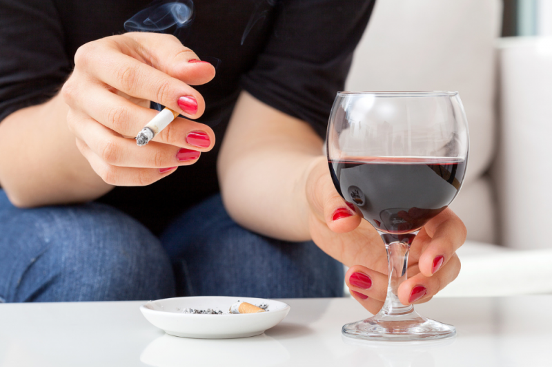 A hand reaches for a glass of red wine on a table, while the other hand holds a cigarette