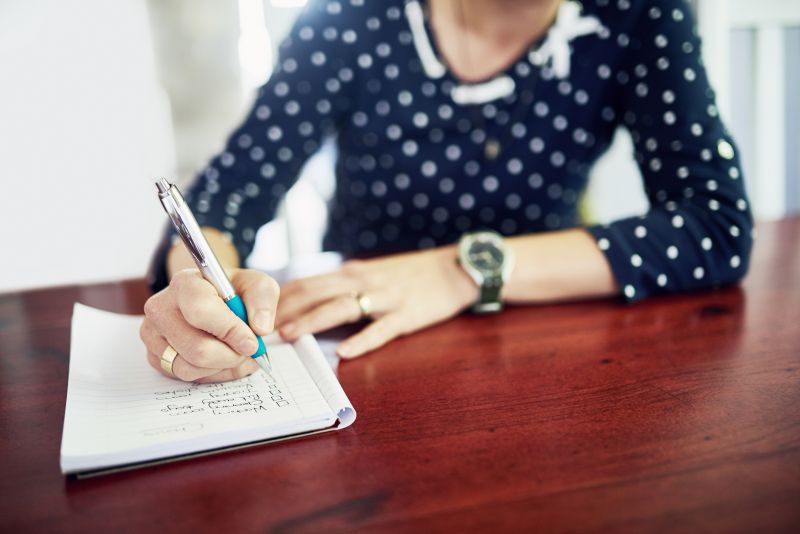 A woman's hand is shown holding a pen, writing a 'to do' list in a notebook.