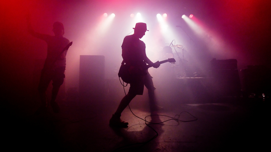 A guitarist on stage playing a solo. He is wearing a hat and is backlit by pink and white lights