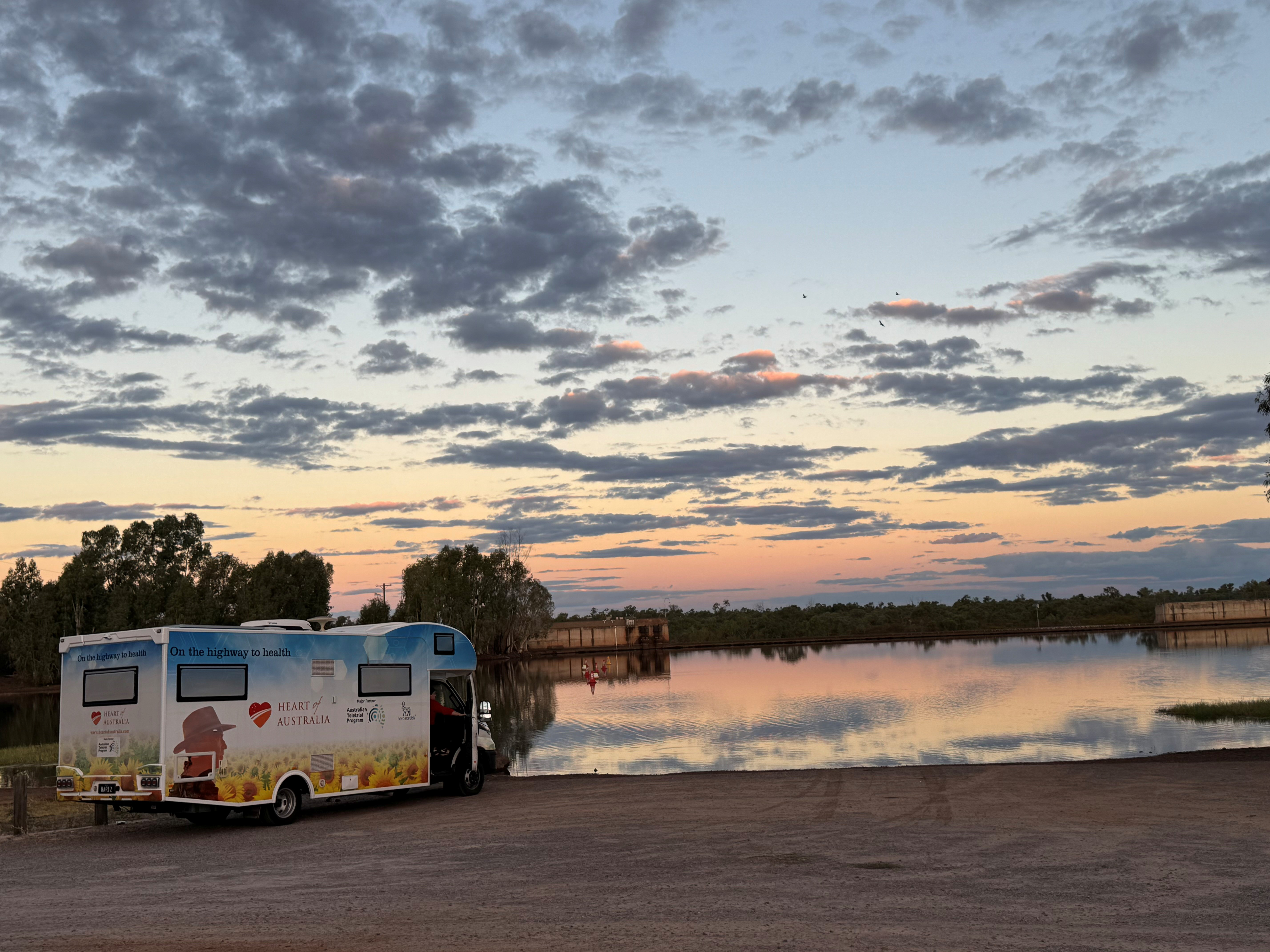 The white Heart of Australia van is parked in front of a body of water as the sun sets behind it.