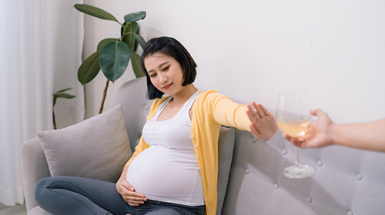 A young pregnant woman on a couch holds up her hand to refuse and offered glass of wine