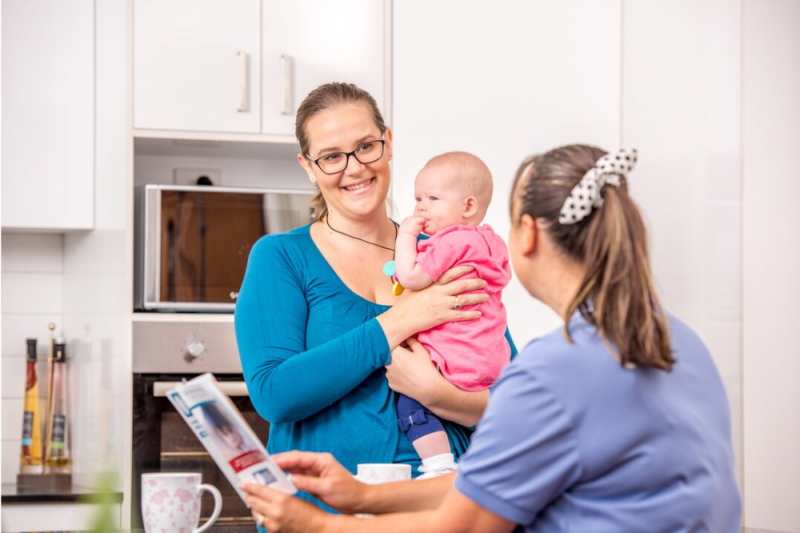 A new mother in her kitchen holding her baby discusses the baby's nutrition with a Mums and Bubs nurse