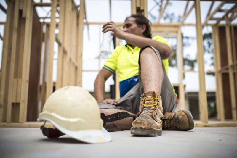 A male construction worker takes a break, sitting against a building frame and drinking water.