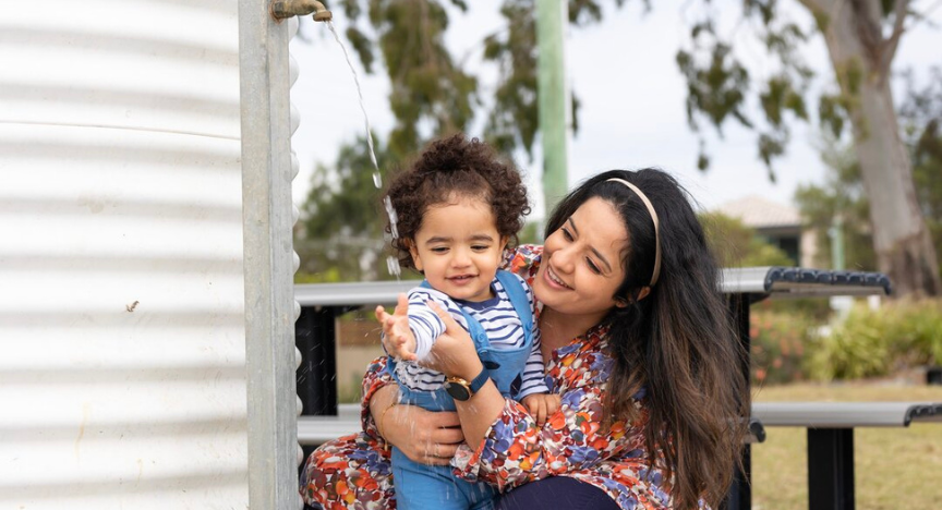 A woman kneels and holds her toddler while the child reaches out for a stream of water from a water tank tap