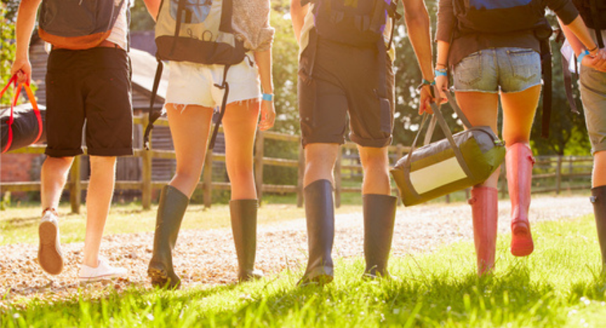 A group of people in gumboots walk into a music festival, lit by afternoon sun.