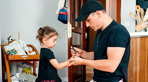 A kneeling father does a finger prick blood sugar test on his young daughter who has type 1 diabetes