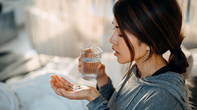 A young Asian woman with antibiotic capsules in her left hand and a glass of water in her right. She is looking at the capsules.