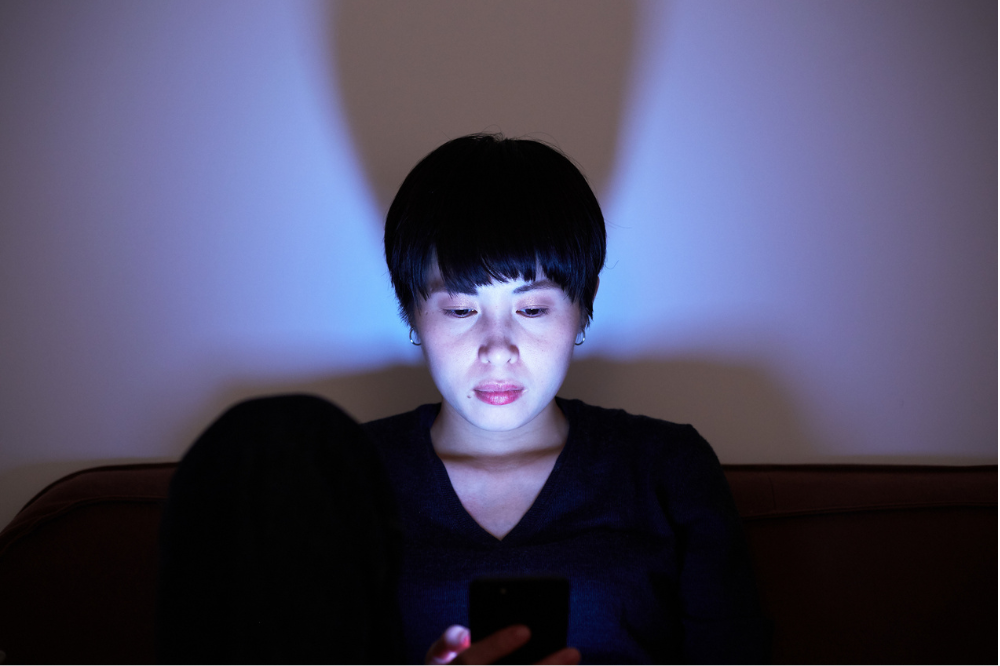 A women sits in a darkened room, her face highlighted by blue light from a digital device in her hands