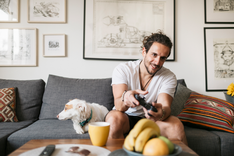 A man with healthy snacks playing games on a couch alongside his dog