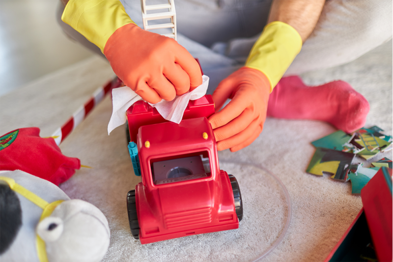 A person wearing orange and yellow rubber gloves cleans a red toy truck with wipes