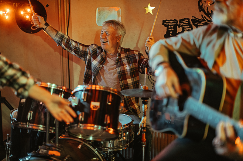 A laughing mature man playing drums in a band with his friends acknowledges the audience