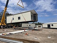 You can see a modular unit being lowered by a crane into place on site at Biggenden. Construction workers monitor the crane-in of the unit. You can see the front and one side of the modular unit. There is construction materials in the foreground.