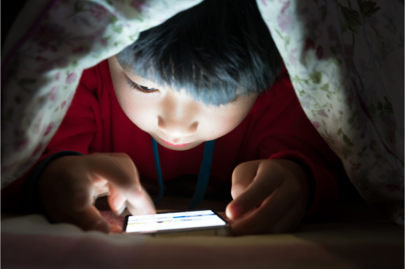 A young child is under the bedcovers in the dark looking at a mobile phone with the light from the phone lighting up his face