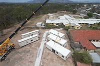 You can see a crane to the left of the image setting a fifth modular unit into place in site. Construction workers unsecure the unit from the crane’s support. There are two modular units on the left hand side and three on the right, separated by a concrete pathway. You can see the Biggenden Multipurpose Health Centre in the background white a white roof. The old nurses quarters sits to the side with the terracotta roof.