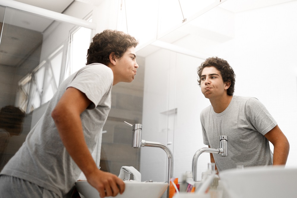 A young man stands at the sink, cheeks puffed out as he rinses his mouth.