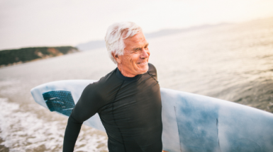 A smiling grey-haired man in a black sun shirt carries a surfboard under his arm at the edge of the ocean