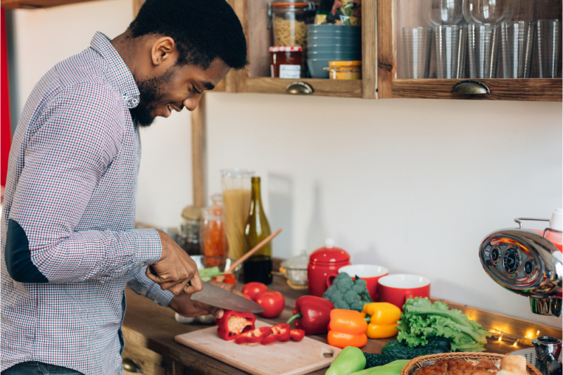 A young African-American man prepares vegetables for a healthy meal in his kitchen. He is chopping a red pepper.