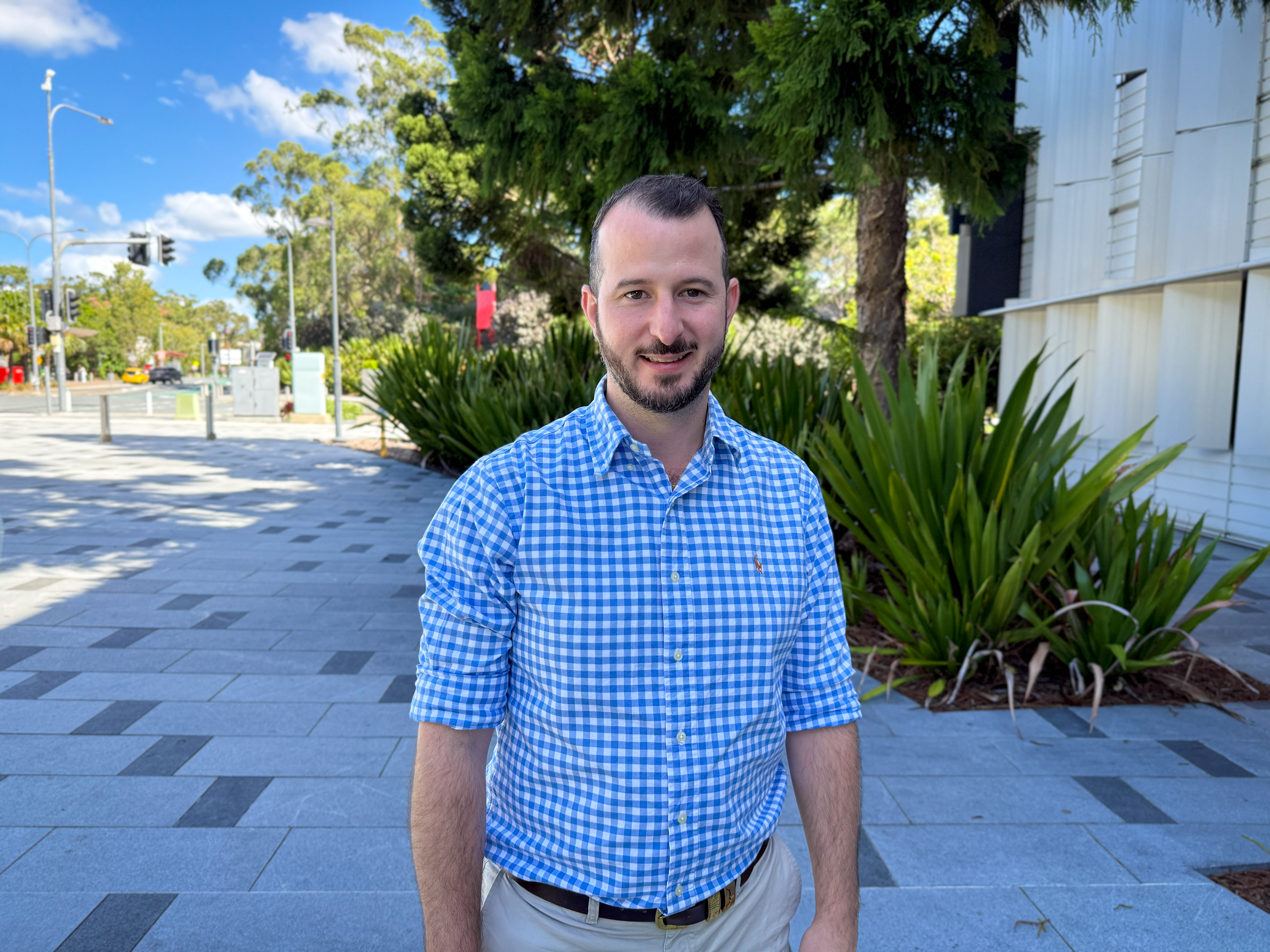 Infectious Diseases Physician Dr Peter Simos. Dr Peter Simos stands straight in a blue shirt and cream pants smiling at the camera with a courtyard in the background.