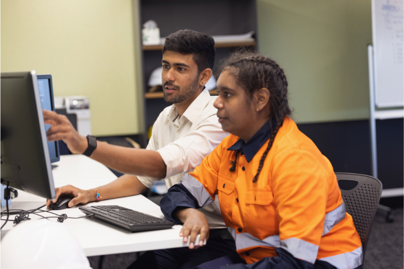 Two office workers, a man and a woman, the latter in a high-vis orange shirt, talk as they sit at a desk looking at the same computer screen.