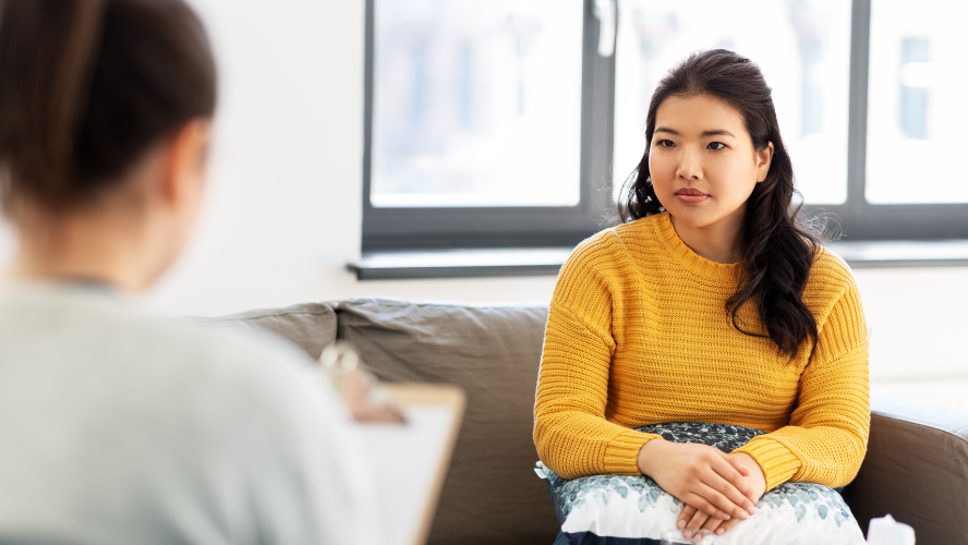 A woman in a yellow jumper sits on a couch while her doctor takes notes
