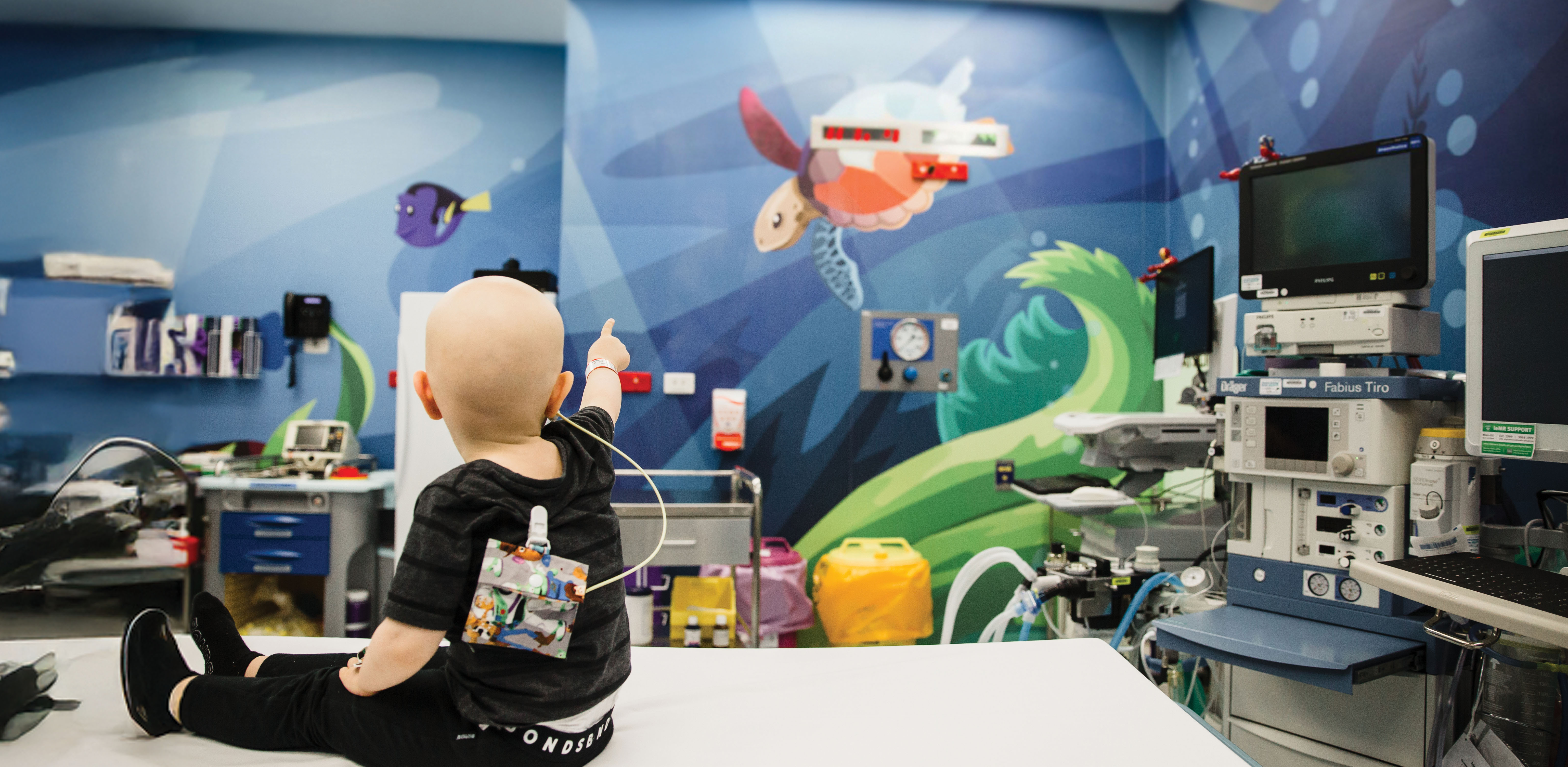 A Hospital patient room featuring a child sitting on a bed and pointing towards a colourful underwater-themed mural, with medical equipment visible in the room.
