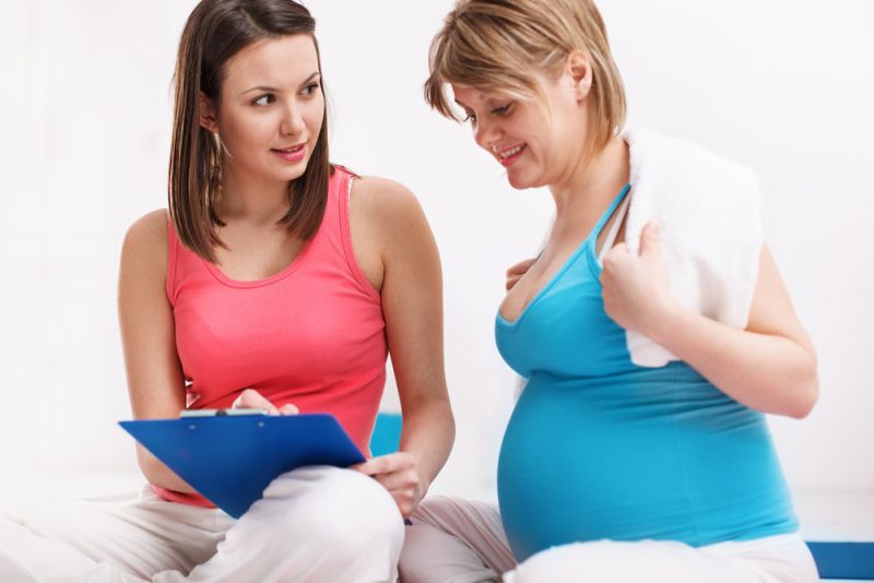 A pregnant woman sits with her physio on an exercise mat, looking over her exercise plan.