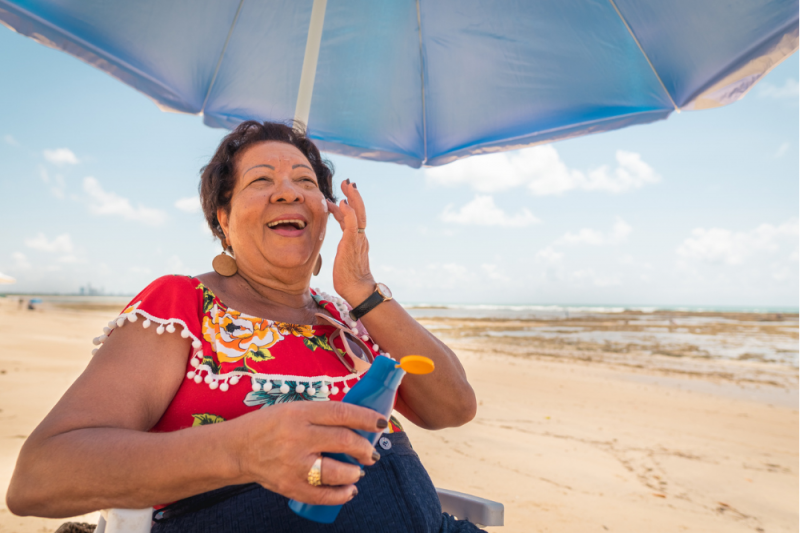 A smiling, mature woman in a bright red dress sits on the beach under an umbrella, applying sunscreen to her cheek