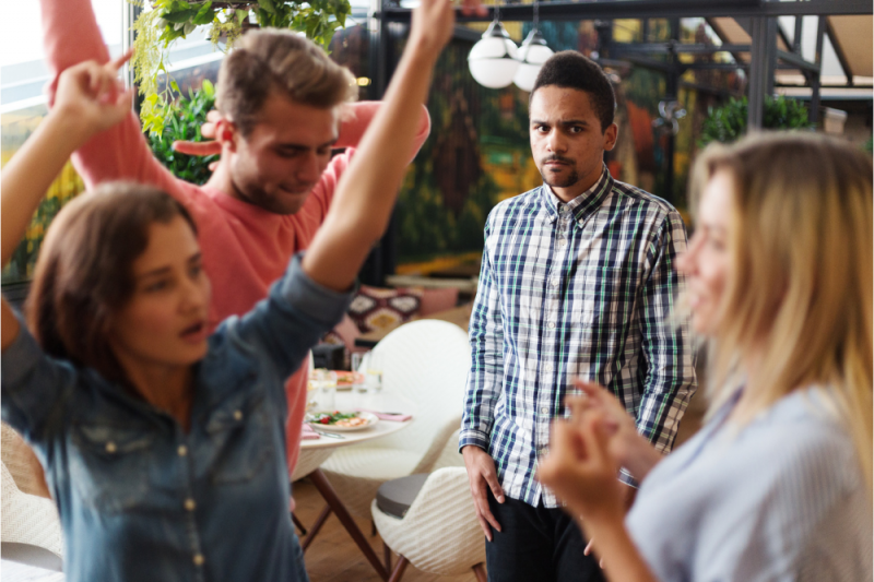 A young man at a loud, bright party looks overwhelmed and anxious while watching people dance