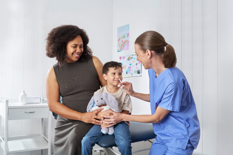 Child seated with a parent while a healthcare worker gives a nasal spray vaccine in a clinic.