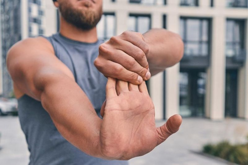 An athletic young man with a beard dressed in a grey singlet stretches the palm of his hand by extending his arm straight in front of him and pulling back on his fingers with the other hand