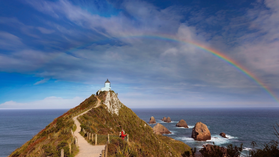 A path winds up to a lighthouse on a cape. Two hikers stare at a rainbow over the sea
