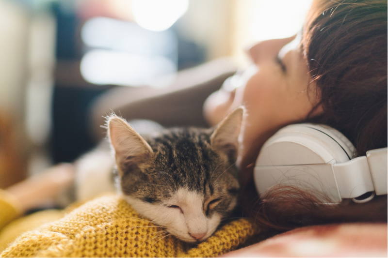 A young woman in a soft mustard sweater with headphones on relaxes back with her young cat lying across her 