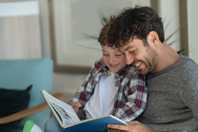 A man reading to his child on the couch. They both are smiling and look slightly shy