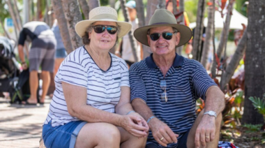 A smiling hat-wearing senior couple sitting close together in the shade on the wall at Southbank