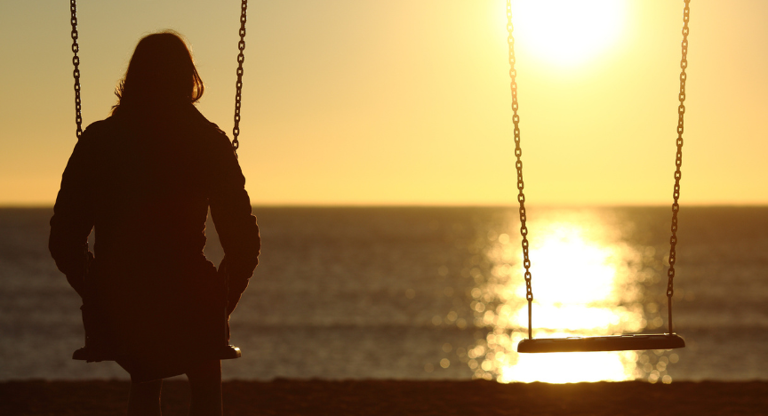 A woman sits on one of two swings staring out at the sunrise over the ocean