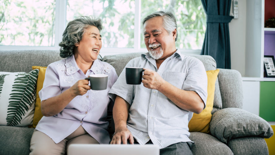 A couple sit on the couch and cheers their mugs.