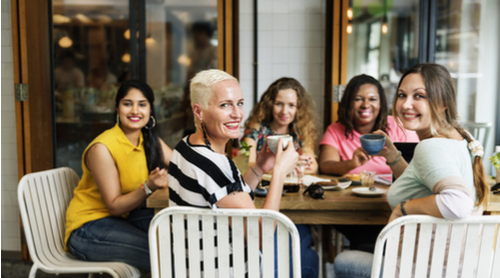 A group of women of different ages sit around a large table, drinking coffees and smiling at the camera.