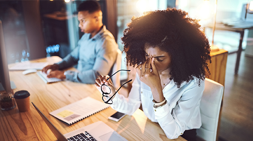 A woman sitting in front of a computer has removed her glasses and is rubbing her tired eyes. A male colleague sits along the table from her.