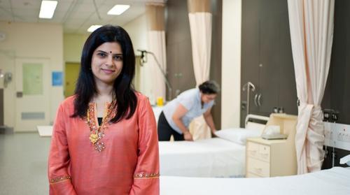 A woman in a red dress stands in a hospital ward in front of a row of beds. In the background a hospital worker is making one of the beds