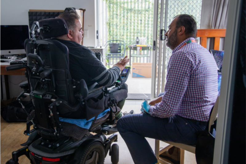 Patient Sam is in a wheelchair in his home sitting and chatting with nurse navigator Dan
