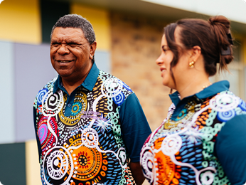 Two individuals in vibrant shirts stand outdoors, smiling and enjoying the sunny weather.