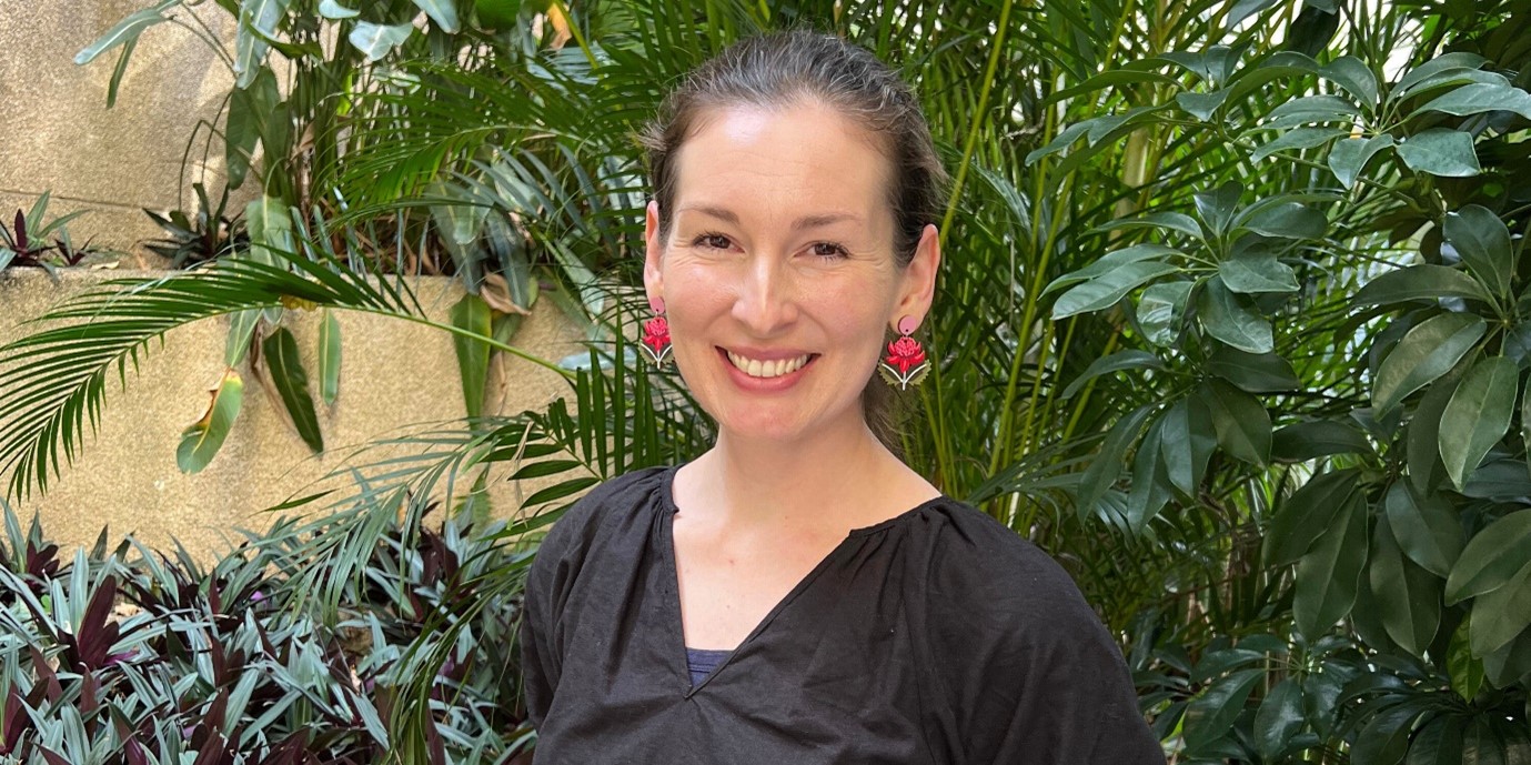 Woman with black blouse and red flower earrings stands in front of a green garden backdrop