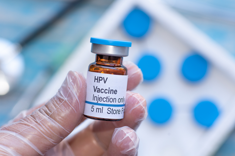 A latex gloved hand holds a vial of HPV vaccine