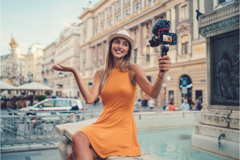 A social media influencer with a professional camera records herself sitting on a fountain in Italy