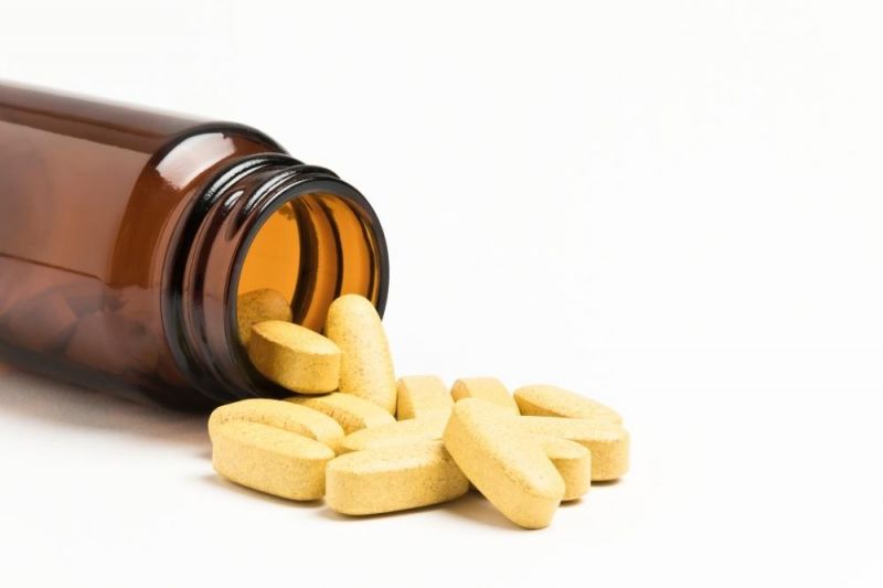 An open brown glass bottle of vitamins lies on it's side spilling large pale yellow tablets onto a white surface