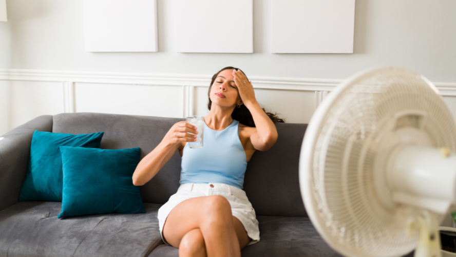 A woman sits on a couch with a glass of water and a fan